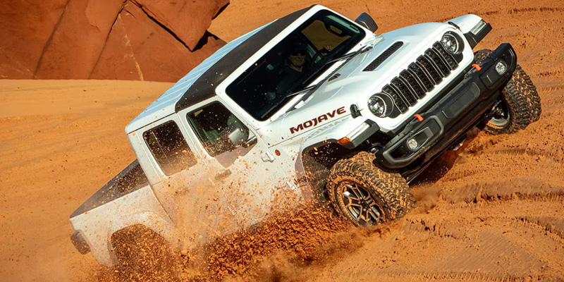 A white Jeep Gladiator Mojave driving through sandy terrain, kicking up dust.